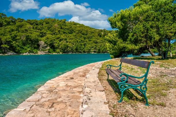 A rustic wooden and cast iron bench sits empty beside a stone walking path on St. Mary's Island in beautiful Veliko Jezero Lake, in Mljet National Park, Croatia.