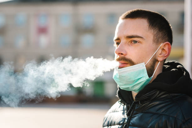 coronavirus. fumar. hombre primer plano con máscara durante la pandemia covid-19 fumando un cigarrillo en la calle. fumar causa cáncer de pulmón y otras enfermedades. los peligros y el daño de fumar. - anti cancer fotografías e imágenes de stock