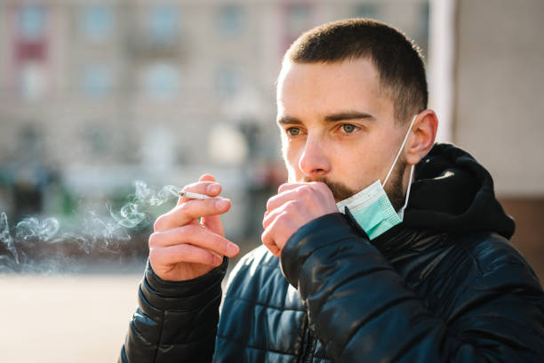 coronavirus. smoking. close up man with mask during covid-19 pandemic coughing and smoking a cigarette at the street. smoking causes lung cancer and other diseases. the dangers of smoking. - nicotine healthcare and medicine smoking issues lifestyles imagens e fotografias de stock