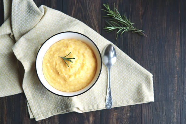 Creamy corn traditional polenta or mamaliga with butter. Served in white shallow dish on rustic wooden table with rosemary. Creamy corn traditional polenta or mamaliga with butter. Served in white shallow dish on rustic wooden table with rosemary. Country style. Selective focus, copy space, top view. Healthy food concept mamaliga stock pictures, royalty-free photos & images