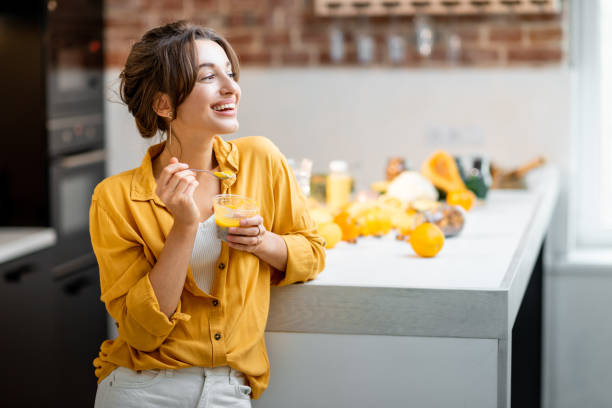 Woman eating chia pudding in the kitchen Portrait of a young and cheerful woman eating chia pudding, having a snack or breakfast in the kitchen with lots of fresh fruits and vegetables. Concept of vegetarianism, healthy eating and wellness eating chia seeds stock pictures, royalty-free photos & images
