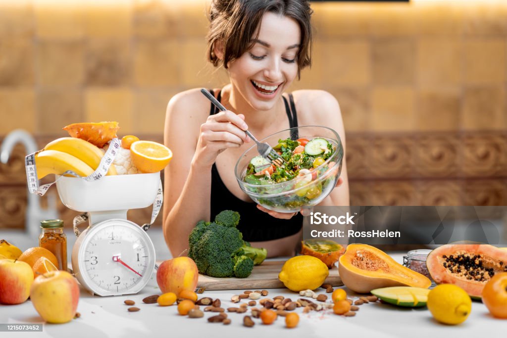 Sports woman with healthy food on the kitchen Sports woman eating salad, standing with lots of healthy fresh food on the kitchen. Concept of losing weight, sports and healthy eating Healthy Eating Stock Photo