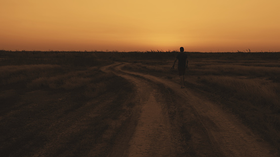 A man in shorts and a T-shirt is walking along a dirt road. Wide angle sunset