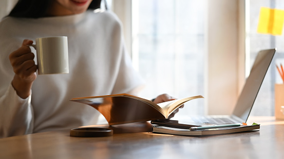 Cropped image of beautiful woman holding a coffee cup in hand while reading a book and sitting in front her computer laptop that putting on wooden table over comfortable living room as background.