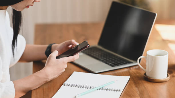 istock Cropped image of beautiful woman working as secretary holding/using a black blank screen smartphone while sitting in front her blank screen computer laptop over comfortable living room as background. 1215075991