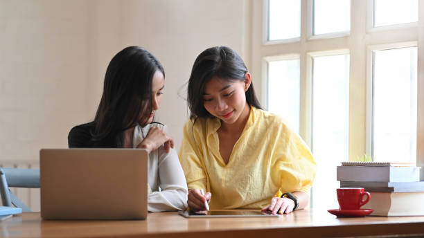 istock Young women talking together while sitting at the wooden working table with computer laptop and tablet next to living room windows as background. 1215076060