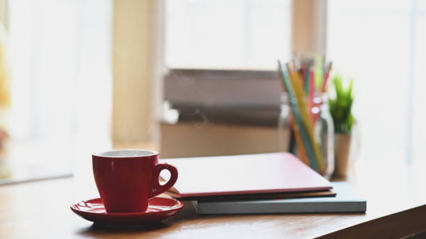 istock Coffee morning concept, Red coffee cup with coasters putting on wooden table with pencil holder, stack of books, potted plant over living room windows as background. 1215076124