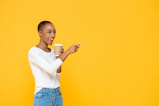 Happy beautiful African American woman drinking coffee smiling and pointing to empty space aside isolated on yellow background