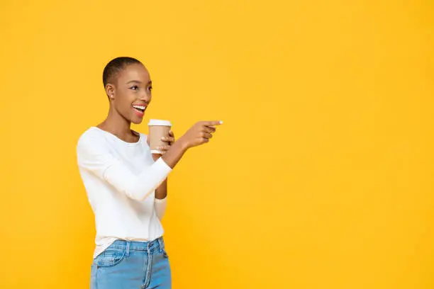 Photo of Happy beautiful African American woman drinking coffee smiling and pointing to empty space aside isolated on yellow background
