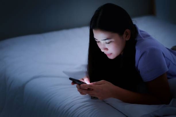 азиатская молодая женщина, лежащая на белой кровати и играющая в смартфоны в ночное время - smart phone asian ethnicity mobile phone women стоковые фото и изображения
