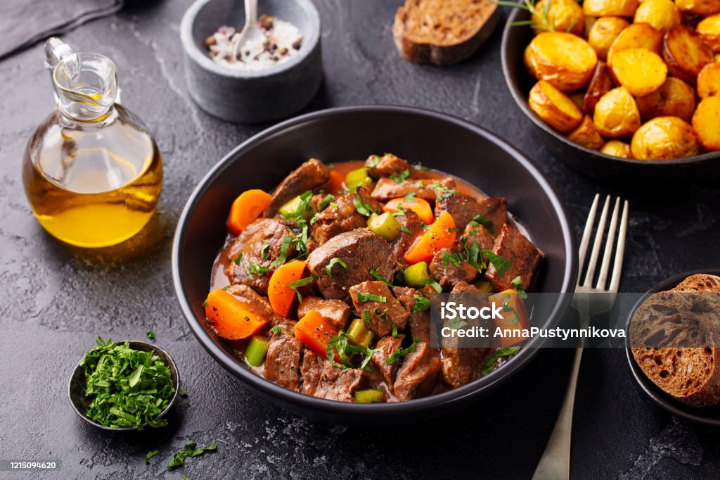 Beef meat and vegetables stew in black bowl with roasted baby potatoes. Dark background. Beef Stew Stock Photo