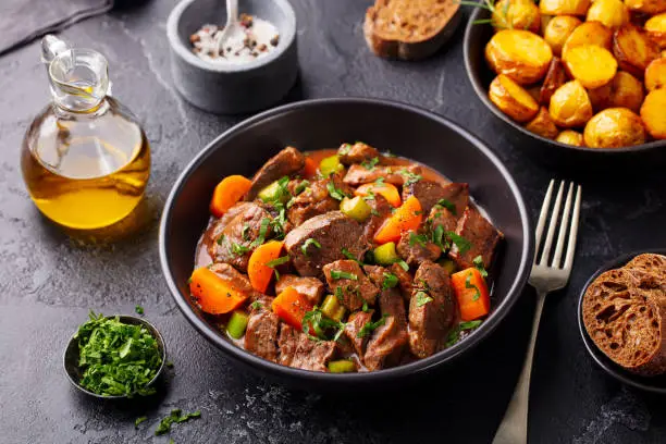 Photo of Beef meat and vegetables stew in black bowl with roasted baby potatoes. Dark background.