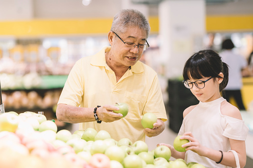 An asian chinese grandfather selecting green apple fruit at the fruit stall with his granddaughter
