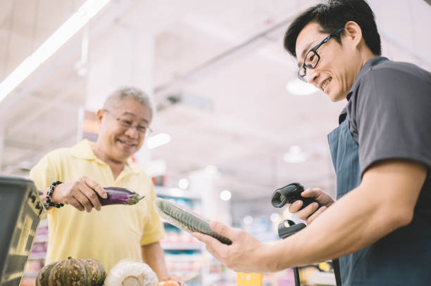 an asian chinese senior adult paying for his shopping items to cashier at counter check out in supermarket - supermarket sales clerk grocer apron imagens e fotografias de stock