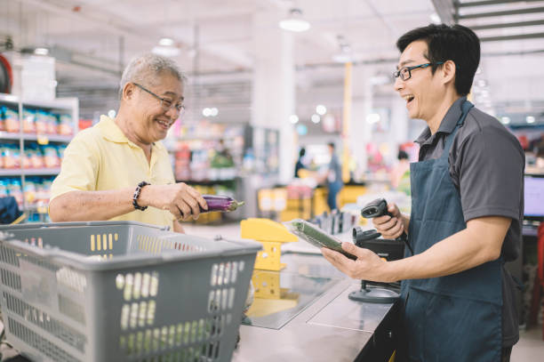 an asian chinese senior adult paying for his shopping items to cashier at counter check out in supermarket - supermarket sales clerk grocer apron imagens e fotografias de stock