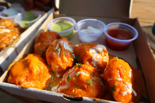 Stock photo showing a cardboard box filled with chicken tikka momos, resembling a traditional chicken tikka masala sauce, complete with spicy tikka sauce and cream / yoghurt curd topping, as well as herbs and spices served with spicy, green and mayonnaise dip. This popular meal is often for sale as street food and is both inexpensive and delicious as an Indian takeaway, being photographed in New Delhi, India. imagen de las madres tikka de pollo / albóndigas caseras en una caja de cartón cubierta con salsa tikka picante grasienta y crema / yogur, hierbas y especias, servido con salsas picantes, comida callejera tradicional comida para llevar india en nueva del