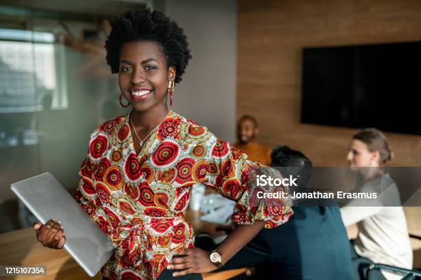 Beautiful Young Smiling Professional Black African Business Woman Holding Laptop Coworkers Hold A Meeting In Background Stock Photo - Download Image Now