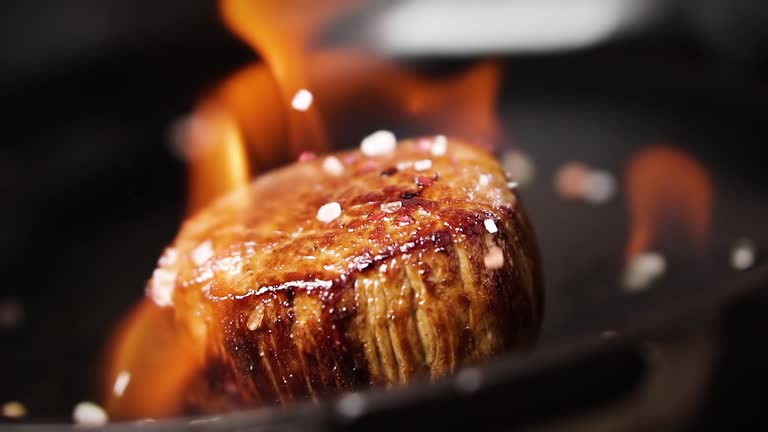 Filet mignon steak is fried in a pan with fire. Salt falls on a steak, slow motion.