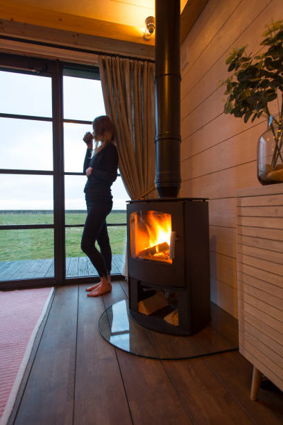 One woman standing next to wood burning stove looking out of the window Single lady standing looking of window at empty garden as wood burning stove roars with flames while sipping a drink from a cup wood burning stove stock pictures, royalty-free photos & images