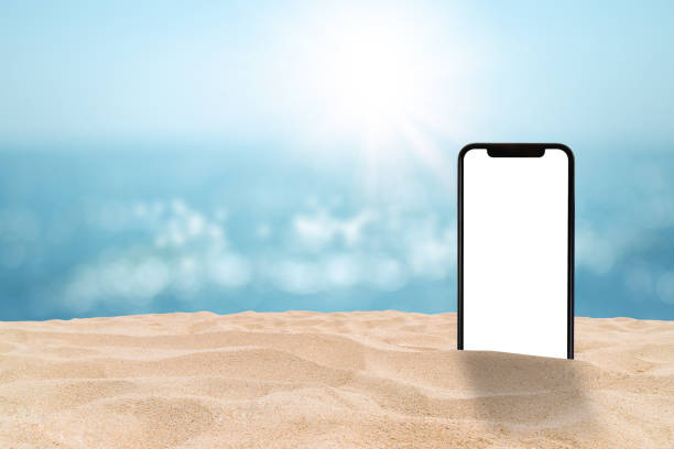 istock Black smartphone with blank white screen on sand beach with blurry seascape view and bokeh sunlight in background. 1215113992