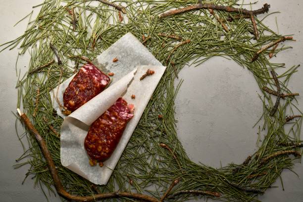 istock Dried sausage from beaver meat on parchment paper and an empty frame from needles. Composition in a frame of branches. 1215114095