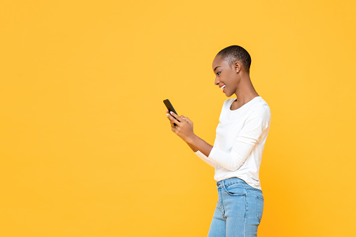 Smiling young African American woman using smartphone  isolated on yellow studio background
