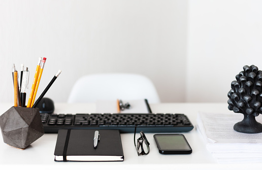 Home office, modern workplace: white table, keyboard laptop computer, concrete holder with pencils and pens, notebook, smartphone, glasses, paperweight with documents, education concept background