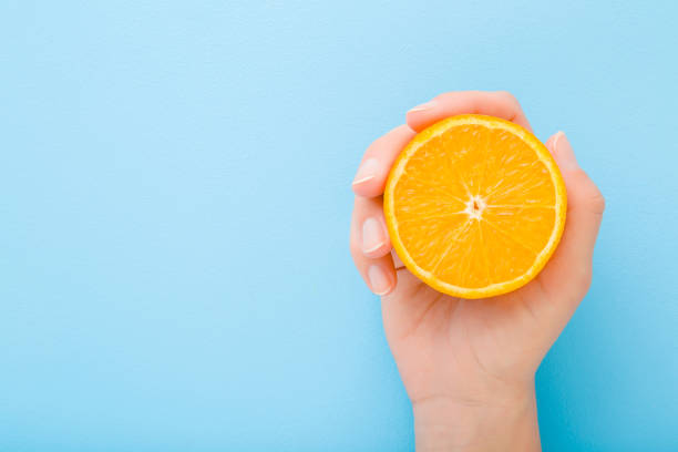 istock Young woman hand holding half of orange. Fresh fruit. Empty place for text on light blue table background. Pastel color. Closeup. Top down view. 1215114949