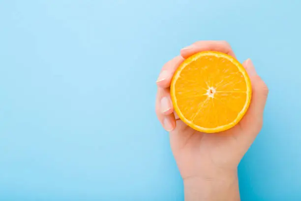Young woman hand holding half of orange. Fresh fruit. Empty place for text on light blue table background. Pastel color. Closeup. Top down view.