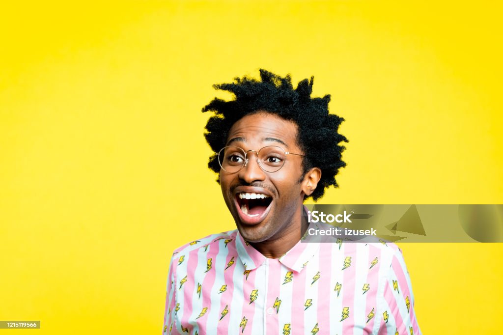 Excited man wearing vintage shirt, portrait on yellow background Summer portrait of happy afro american young man with dreadlocks wearing vintage shirt with lightning pattern and gold glasses, looking away with mouth open. Studio shot on yellow background. Men Stock Photo