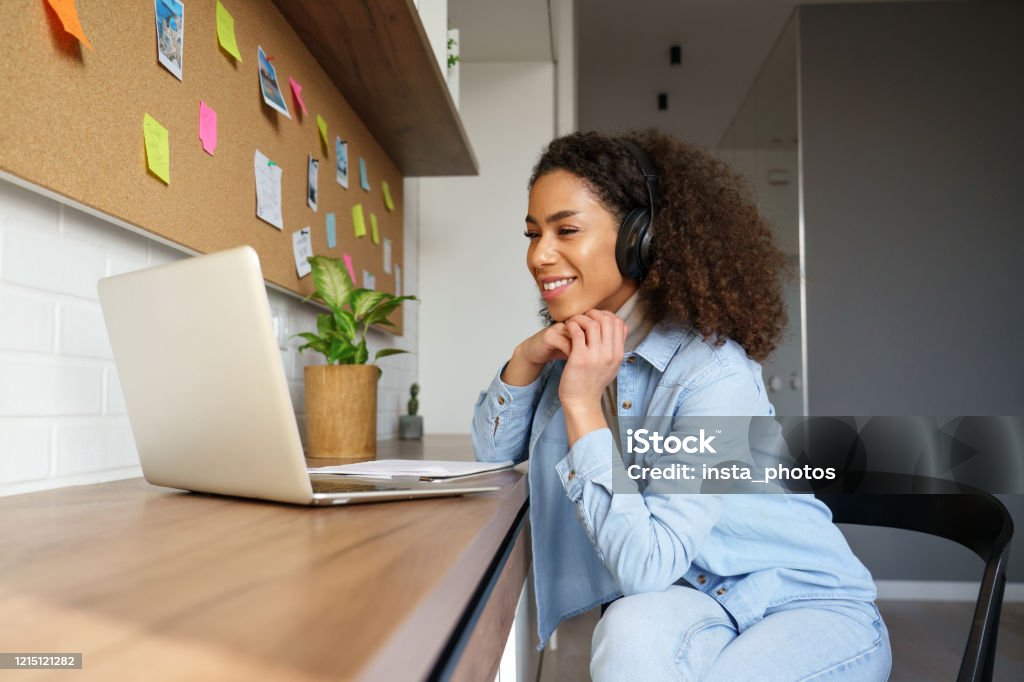 Smiling young african american teen girl wear headphones video calling on laptop. Happy mixed race pretty woman student looking at computer screen watching webinar or doing video chat by webcam. Internet Stock Photo