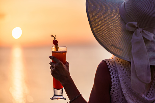 Back view of a woman with a cocktail looking at sunset view at sea. Focus is on hand and cocktail.