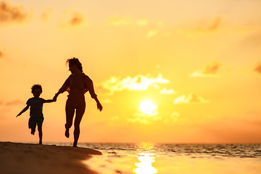 Happy mother and her small son holding hands while running on the beach at sunset. Copy space.