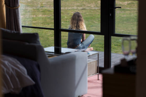 View from inside of house looking out at single woman sat on garden decking in back garden looking at green grass outside