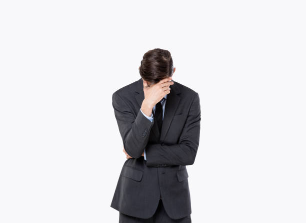 Full length of bored young male entrepreneur with dark hair in formal suit covering face with hand and looking at camera against white background