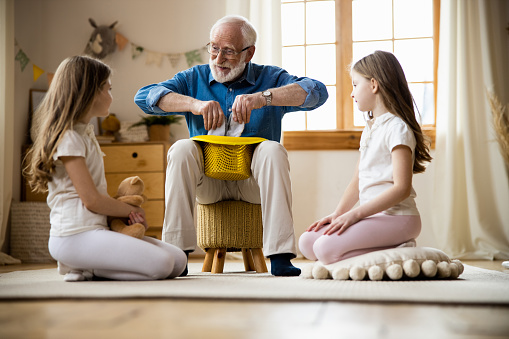Cheerful grandfather pulling toy rabbit by the ears out of a hat and girls watching it