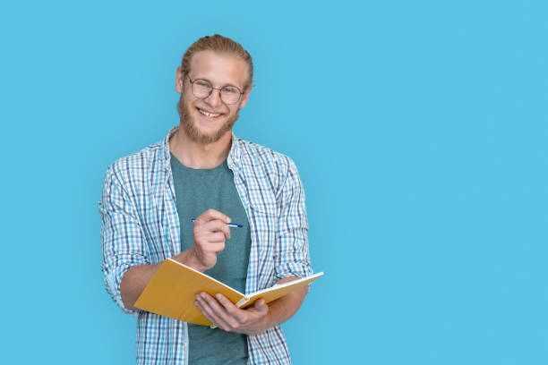 istock Happy young bearded man student hold write in notebook look at camera, casual guy university teacher stand with workbook and pen isolated on blue studio background, portrait, education concept. 1215128929