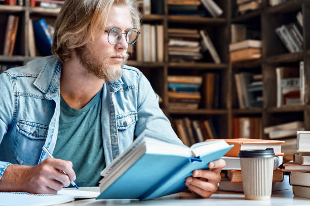 istock Young thoughtful man student researcher glasses look away sit library desk think over task handwrite in notebook make notes from textbook prepare diploma project study for exam test education concept 1215129058