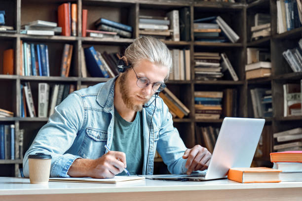 istock Focused young man student wear headset make notes e study online in library listen lecture internet course training online learning foreign language distance education write information in notebook. 1215129077