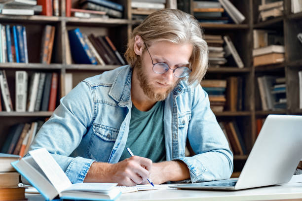 istock Young concentrated male student at library desk with modern laptop pc handwrite in notebook hold pen make notes skype study online training e learning prepare exam distant electronic education concept 1215129283