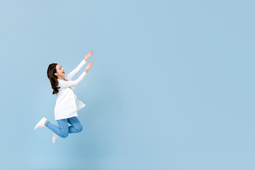 Full length portrait of young happy Asian woman floating in mid-air with hands opening to blank space beside in blue isolated studio background