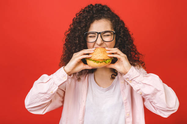 Portrait of young curly beautiful hungry woman eating burger. Isolated portrait of student with fast food over red background. Diet concept. Portrait of young curly beautiful hungry woman eating burger. Isolated portrait of student with fast food over red background. Diet concept. fast food restaurant photos stock pictures, royalty-free photos & images