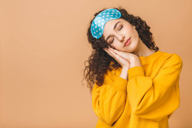Waist up portrait of cheerful young beautiful curly smiling girl in sleeping mask. Isolated on beige background. Waist up portrait of cheerful young beautiful curly smiling girl in sleeping mask. Isolated on beige background. sleep eye mask stock pictures, royalty-free photos & images