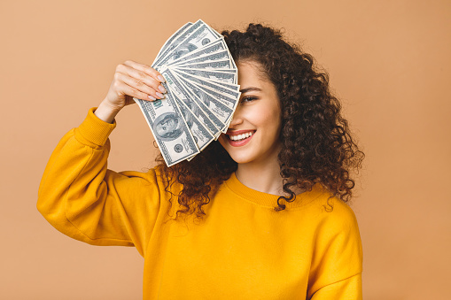 Portrait of a cheerful young woman holding money banknotes and celebrating isolated over beige background.