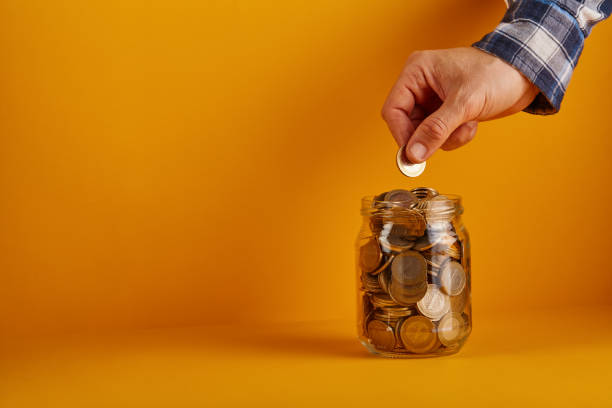 mano di una persona che tiene una moneta da mettere in barattolo di vetro su sfondo giallo / arancione - jar coin currency glass foto e immagini stock