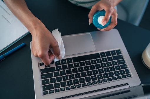 Unrecognizable woman cleans and disinfects her laptop, using disposable wipes and disinfection gel, to prevent Coronavirus