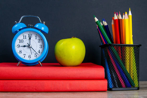 istock The blue alarm clock along with multi-colored pencils stand on a stack of red books, next to an apple 1215152546