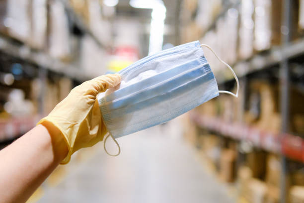 una mano en un guante médico sostiene una máscara en el fondo de un almacén de la tienda. proteger a las personas en lugares públicos de virus y enfermedades, concepto - safety sign protective workwear factory fotografías e imágenes de stock