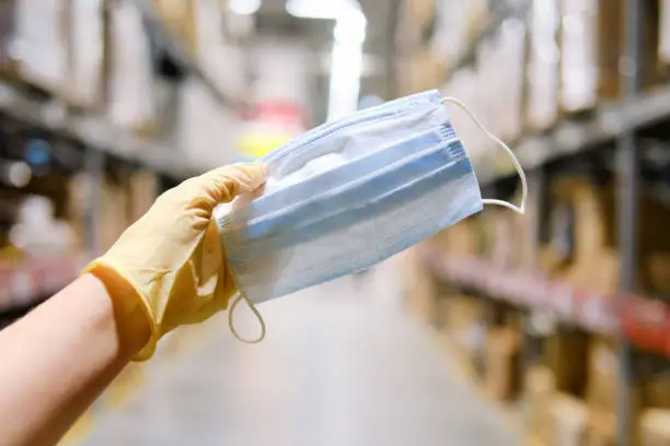 A hand in a medical glove holds a mask against the background of a store warehouse. Protecting people in public places from viruses and diseases, concept