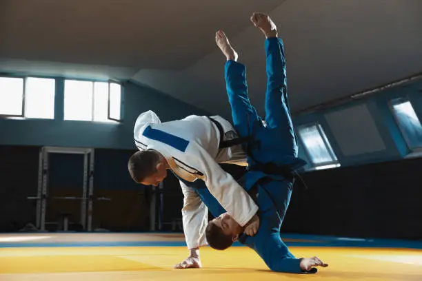 Two young judo caucasian fighters in white and blue kimono with black belts training martial arts in the gym with expression, in action, motion. Practicing fighting skills. Overcoming, reaching target.